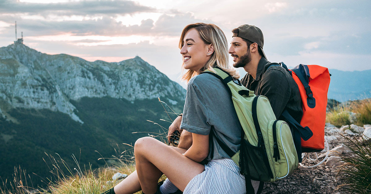 Junges Pärchen sitzt auf einer Wiese in den Bergen und genießt das Panorama. Die junge Frau hat blondes, schulterlanges Haar. Der Mann tägt eine Kappe und einen Vollbart.