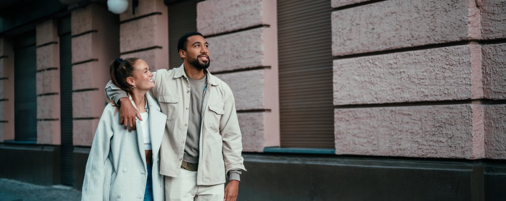 Ein Pärchen spaziert verträumt durch die Stadt. Der Mann hat dunkles Haar und einen dunklen Vollbart, er lächelt und hat den Arm um seine Frau gelegt. Die Junge Frau hat ihr dunkles Haar zu einem Zopf gebunden und lächelt ihren Mann an.